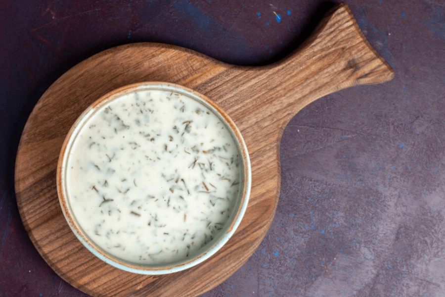 A rustic wooden serving board holds a bowl of creamy white yogurt dip infused with finely chopped herbs. The background features a textured dark purple surface, creating a contrast that highlights the simplicity of the presentation.
