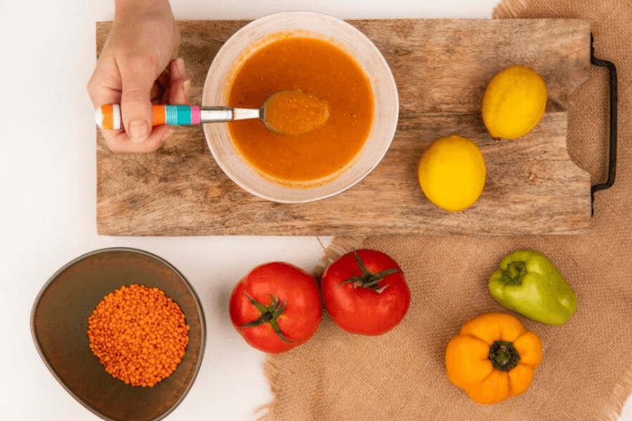 A hand holding a spoonful of orange lentil soup over a bowl on a wooden board, surrounded by fresh vegetables including tomatoes, bell peppers, lemons, and a small bowl of lentils.