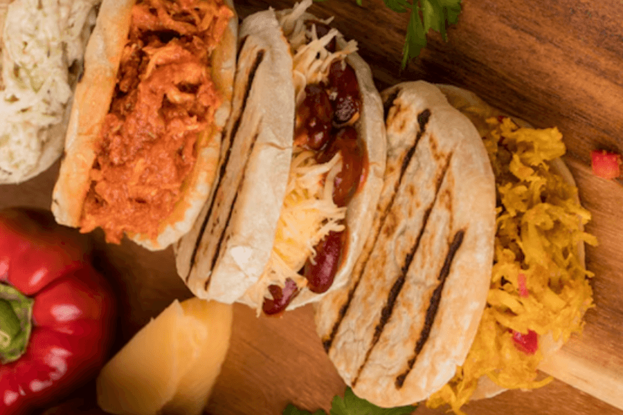Four grilled flatbreads filled with different toppings, including shredded cheese and beans, tomato-based filling, coleslaw, and grated yellow vegetables. They are displayed on a wooden surface with a red bell pepper and parsley as decoration.