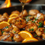 A skillet filled with golden-brown seared chicken bites, garnished with fresh parsley and paired with vibrant orange slices. A drizzle of honey is being poured over the chicken, adding a glossy finish. The scene is warmly lit, with a flame flickering in the background, enhancing the dish's rich and flavorful appeal.