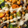 Roasted vegetables with chickpeas, broccoli, Brussels sprouts, and sweet potatoes drizzled with creamy dressing in a white bowl.