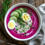 Overhead photo of cold beet soup garnished with dill, cucumber, and boiled egg in a white bowl