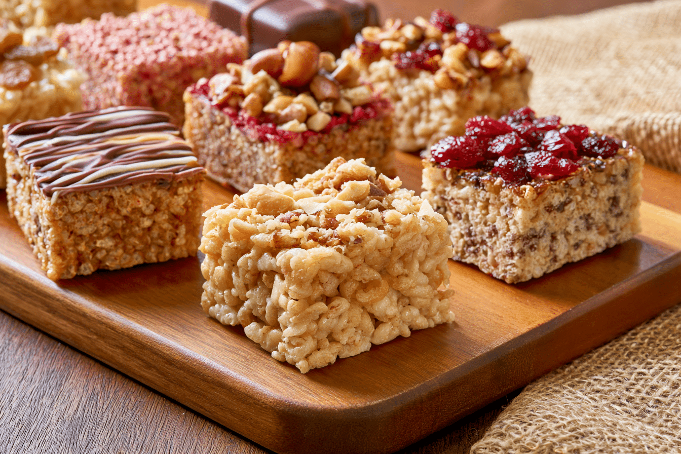 A variety of customized crispy treats on a wooden board, showcasing different toppings like chocolate drizzle, nuts, and dried fruit, with some treats made with alternative grains.